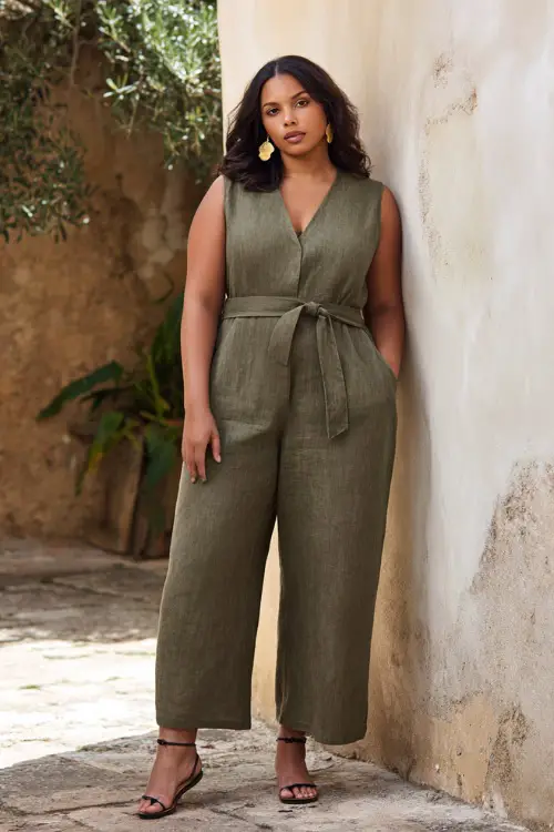 A plus-size woman wears a loose linen jumpsuit with a cinched waist and flattering structure, styled with strappy sandals and statement earrings