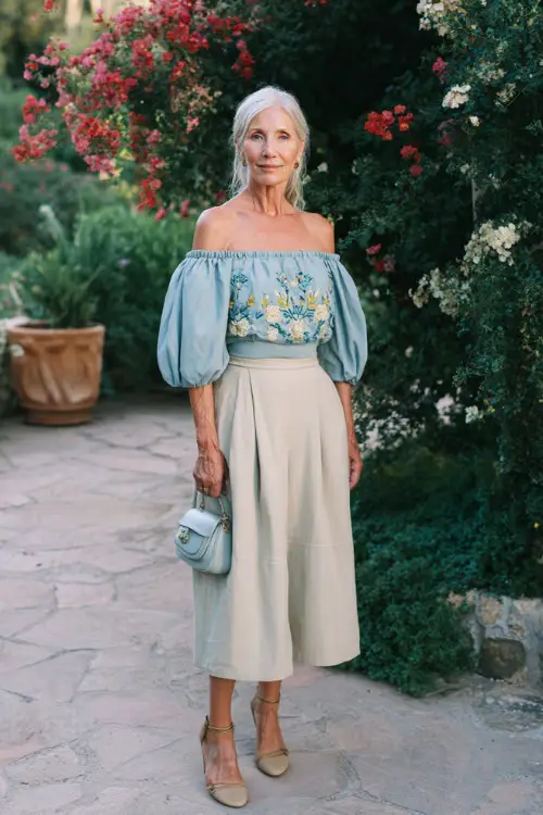 A woman over 50 wears a chic off-shoulder blouse with embroidery paired with a high-waisted midi skirt 