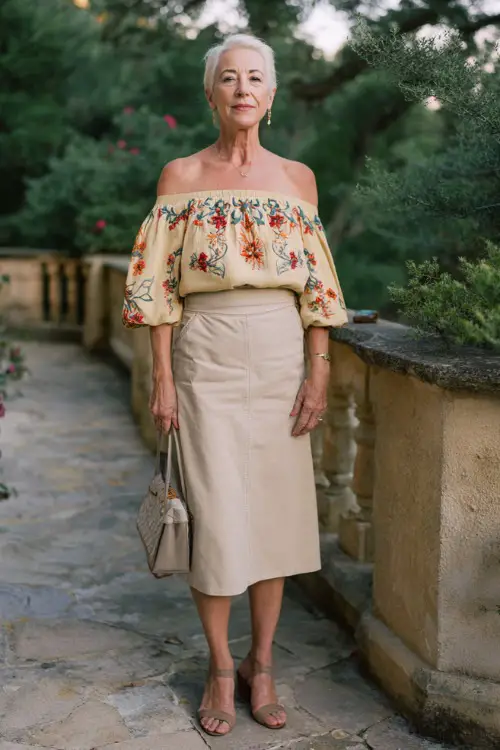 A woman over 60 wears an off-shoulder blouse with embroidery paired with a high-waisted midi skirt 