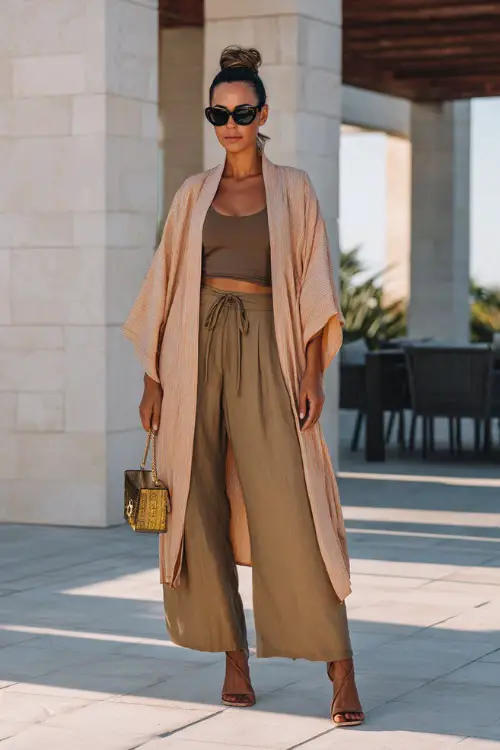 A woman wears a lightweight kimono layered over a fitted top and tailored wide-leg pants, styled with heeled sandals and a structured bag