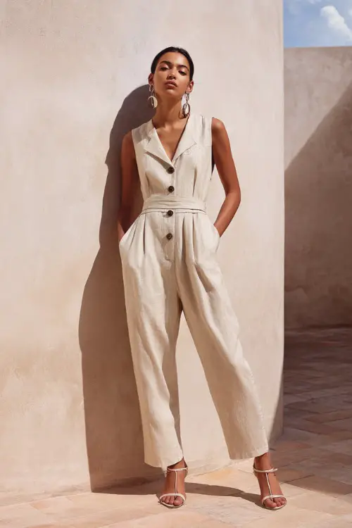 A woman wears a linen jumpsuit with a cinched waist and elegant structure, styled with strappy heels and statement earrings