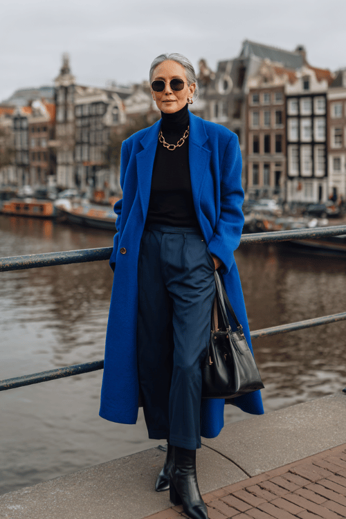 Elegant monochrome outfit for women over 50 featuring a royal blue long coat layered over a black turtleneck with matching wide-leg trousers