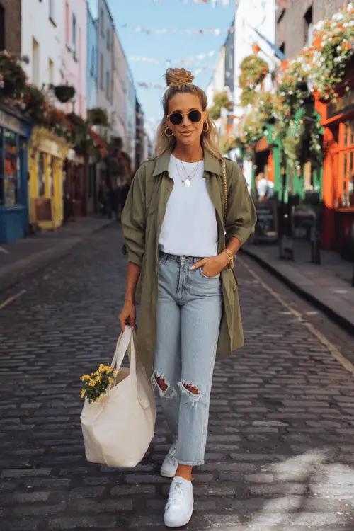 Woman in a casual travel outfit with a white tee, light wash ripped jeans, olive utility jacket, white sneakers, and sunglasses walking down a charming cobblestone city street