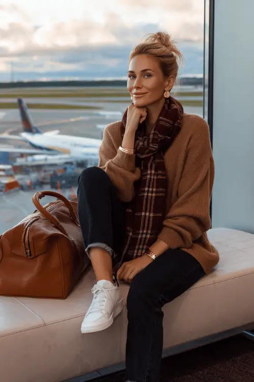 Woman in a cozy airport outfit with a knit sweater, plaid scarf, jeans, white sneakers, and leather duffel bag sitting by the terminal window