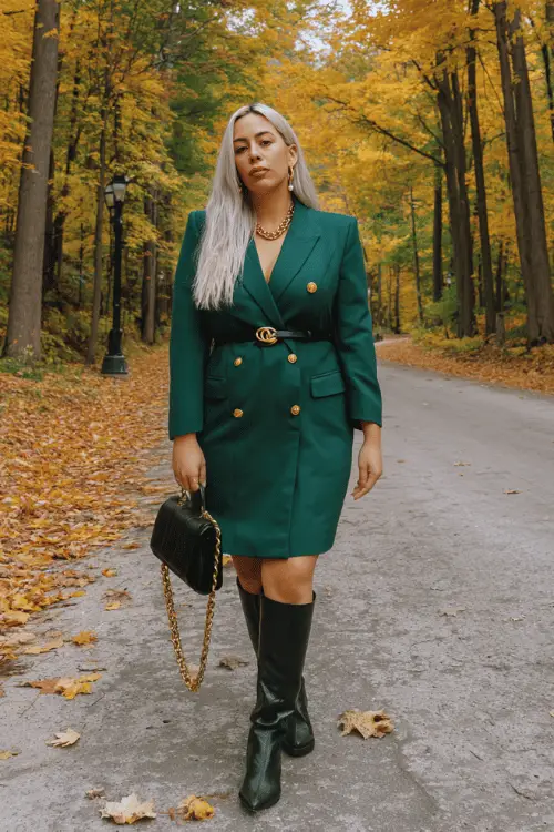 Woman wearing a dark green belted coat dress with black ankle boots, sunglasses, and a chain shoulder bag, posing on a tree-lined path surrounded by autumn foliage 