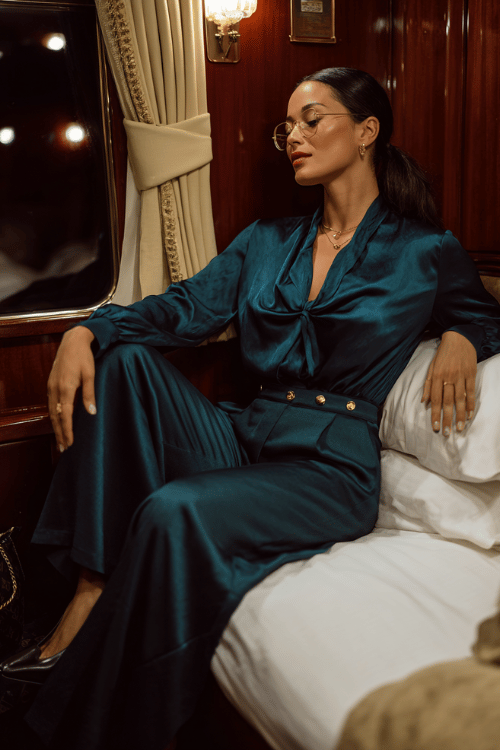 Woman wearing a dark teal satin blouse and matching wide-leg pants relaxing inside a luxury train cabin at night