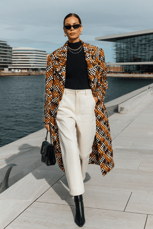 Woman wearing a patterned long coat, black turtleneck, cream wide-leg pants, and black ankle boots walking by a modern waterfront