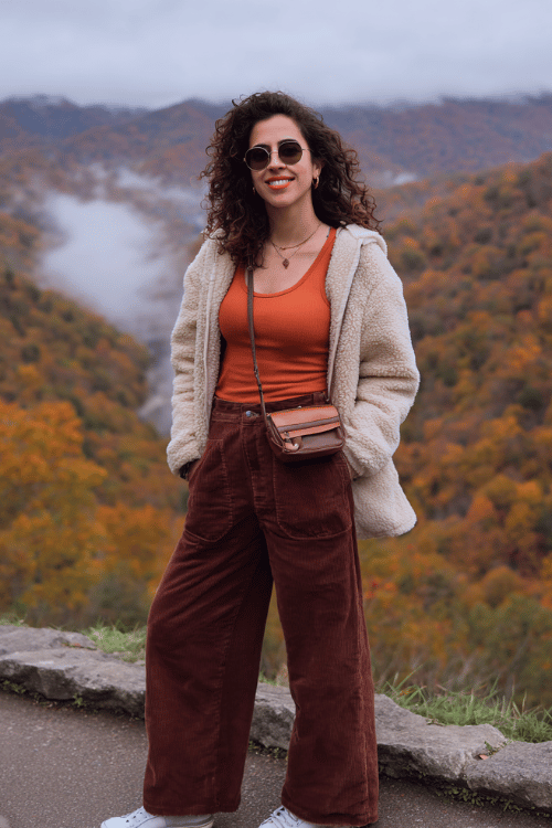 Woman wearing a rust-orange tank top, brown corduroy wide-leg pants, and a cream sherpa jacket at a scenic autumn mountain overlook