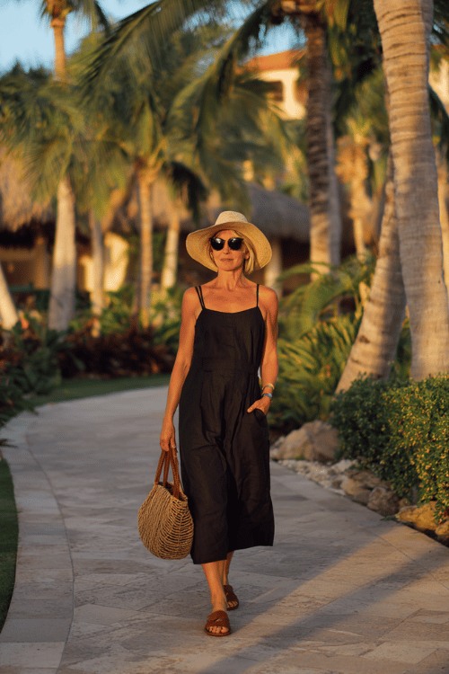 woman wearing a sleeveless black flowy midi dress with flat brown leather sandals, oversized sunglasses, a straw sun hat, and a textured woven handbag