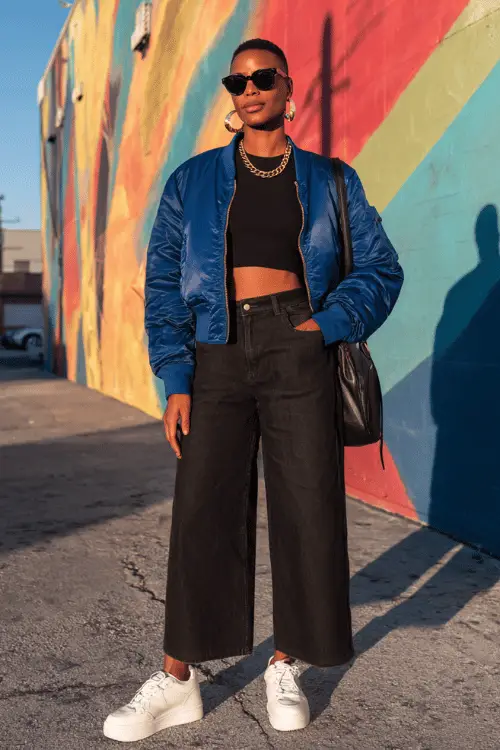 woman with short textured natural black hair wearing a smart casual travel outfit featuring a fitted black long-sleeve henley top tucked into high-waisted wide-leg dark wash denim jeans 