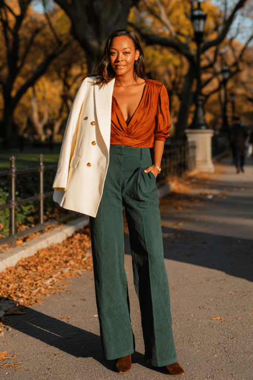 a Black midsize woman wearing a bold burnt sienna wrap blouse tucked into tailored wide-leg trousers in deep forest green, layered with a fitted ivory double-breasted blazer 