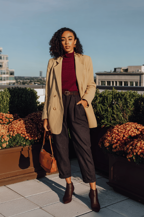 a Black woman wearing an oversized camel blazer belted at the waist over a deep burgundy fitted turtleneck and straight-leg dark wash trousers, finished with pointed-toe ankle boots 