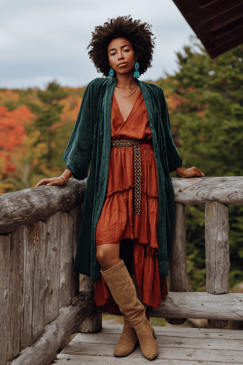a Black woman with a bold afro wearing a deep olive green velvet kimono jacket over a tiered rust-orange chiffon midi dress