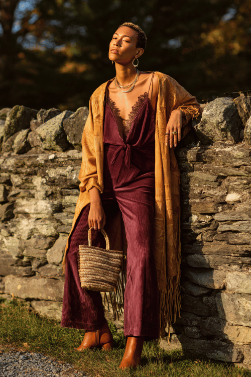 a Black woman with short natural hair wearing a plum velvet flare-leg jumpsuit with lace trim details, draped in a long fringed kimono in burnt sienna and mustard