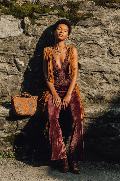a Black woman with short natural hair wearing a plum velvet flare-leg jumpsuit with lace trim details, draped in a long fringed kimono in burnt sienna and mustard