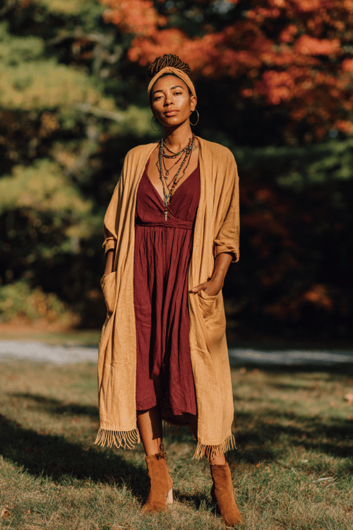 a Black woman with shoulder-length locs wearing a flowing camel-toned duster coat with fringe detailing over a rich burgundy wrap dress