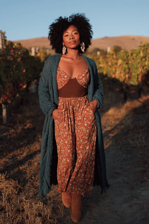 a Black woman with voluminous curly hair wearing a romantic sheer floral maxi dress in warm spice tones over a fitted chocolate brown bodysuit, layered with a long open-knit cardigan in deep teal