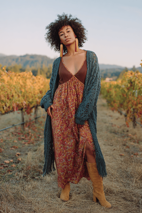 a Black woman with voluminous curly hair wearing a romantic sheer floral maxi dress in warm spice tones over a fitted chocolate brown bodysuit, layered with a long open-knit cardigan in deep teal