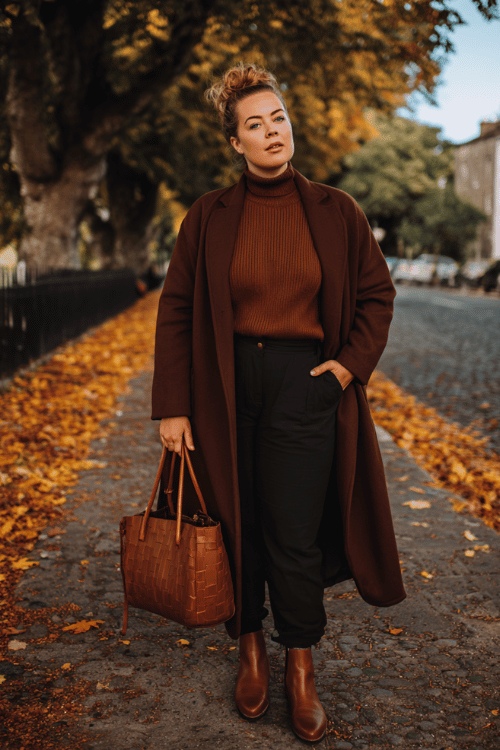 a beautiful plus-size woman wearing a longline wool overcoat in rich deep camel over a fitted ribbed turtleneck in warm oatmeal and tailored straight-leg dark trousers