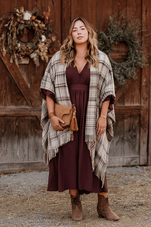 a curvy model wearing a cozy oversized earth-tone plaid shawl draped over a deep wine-colored smocked midi dress with flutter sleeves, paired with stacked wooden bangles