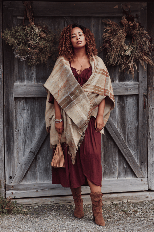 a curvy model wearing a cozy oversized earth-tone plaid shawl draped over a deep wine-colored smocked midi dress with flutter sleeves, paired with stacked wooden bangles