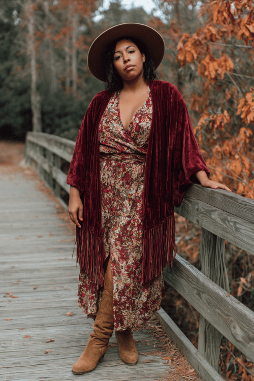 a curvy woman dressed in a deep burgundy velvet kimono jacket over a floral printed wrap dress, wide-brim felt hat, and brown suede fringe boots