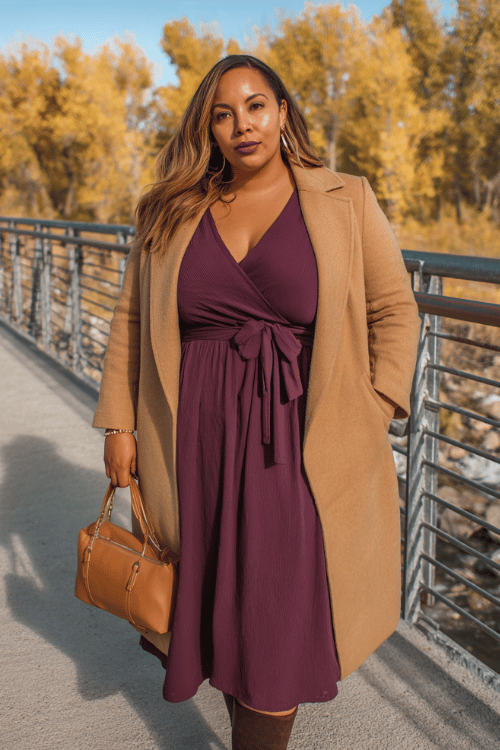 a plus-size woman in a deep plum long-sleeve wrap midi dress with a flattering defined waist and flowing skirt, layered with a longline camel overcoat with structured lapels