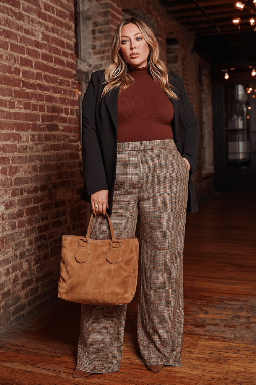 a plus-size woman in a rich mahogany ribbed turtleneck tucked into wide-leg trousers in a bold rust and camel plaid print, layered with a longline black blazer with structured shoulders