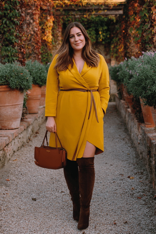 a plus-size woman in a rich saffron yellow longline blazer dress with a cinched waist and asymmetric hem, paired with over-the-knee boots in dark chocolate suede and a structured top-handle bag 