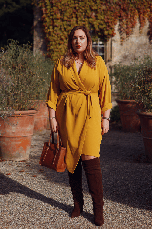 a plus-size woman in a rich saffron yellow longline blazer dress with a cinched waist and asymmetric hem, paired with over-the-knee boots in dark chocolate suede and a structured top-handle bag