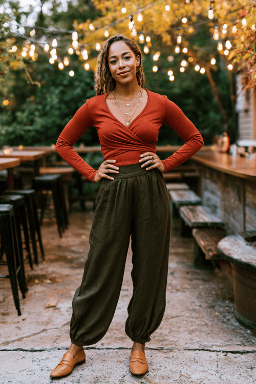 a stylish woman wearing a burnt orange long-sleeve wrap top, wide-leg dark olive trousers, and tan loafers, with layered dainty gold necklaces
