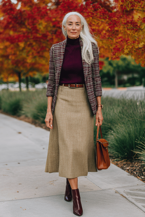 a white-haired graceful woman wearing a deep wine-colored cashmere turtleneck tucked into a high-waisted A-line midi skirt in rich caramel tweed