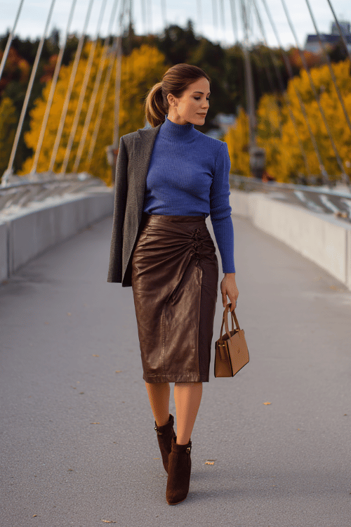 a woman dressed in a soft cobalt blue mock-neck sweater tucked into a high-waisted wrap midi skirt in warm chocolate brown leather-look fabric, layered with a fitted charcoal grey blazer