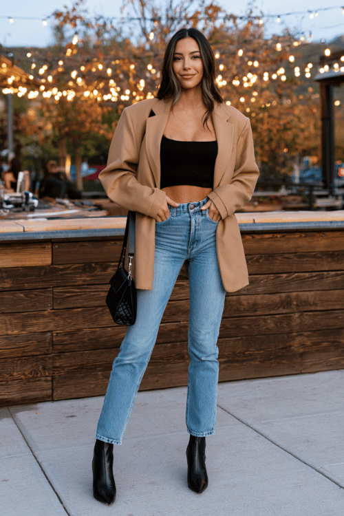 a woman dressed in an oversized camel blazer layered over a black fitted crop top, straight-leg medium wash jeans, and black heeled ankle boots, carrying a small black quilted bag