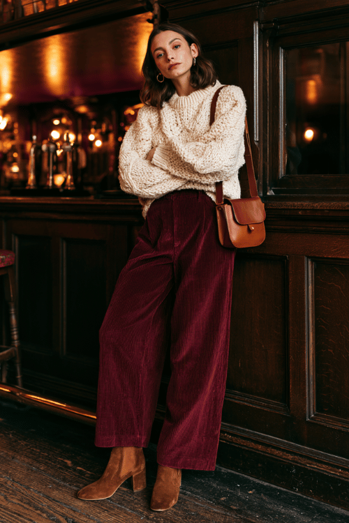 a woman in a chunky knit cream sweater, high-waisted burgundy corduroy wide-leg pants, and brown suede block-heeled boots, carrying a small brown leather shoulder bag