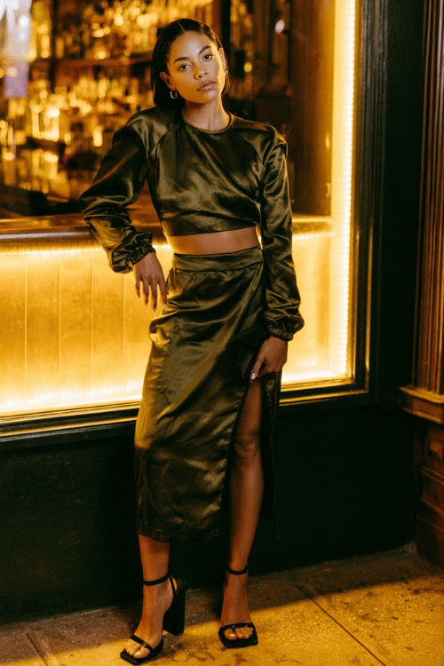 a woman in a dark olive satin two-piece set a cropped long-sleeve top and a high-waisted midi skirt with a front slit black block-heeled sandals and a small black clutch