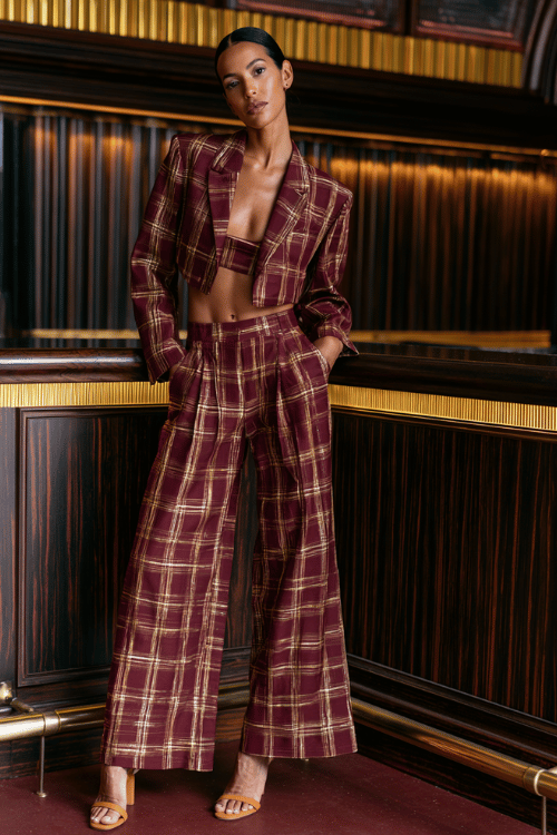 a woman in a plaid two-piece co-ord set in rich burgundy and gold tones, featuring a cropped blazer and matching wide-leg pants, tan strappy heels, posed inside an upscale bar