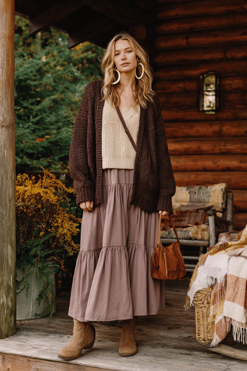 a woman in a relaxed oversized boho pullover in a soft chunky knit of warm cinnamon and cream tones, paired with a flowing tiered skirt in muted dusty mauve 