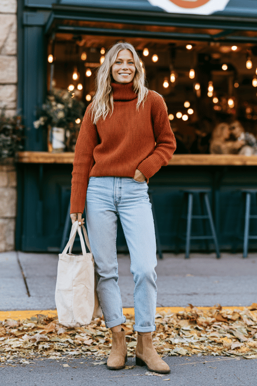a woman in a rust-colored ribbed turtleneck sweater, light wash mom jeans rolled at the ankle, and tan suede Chelsea boots, holding a casual tote bag 
