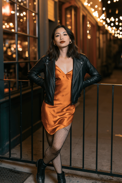 a woman wearing a burnt orange satin bias-cut slip dress layered under a black cropped leather jacket, black fishnet tights, black ankle boots, and small gold hoop earrings