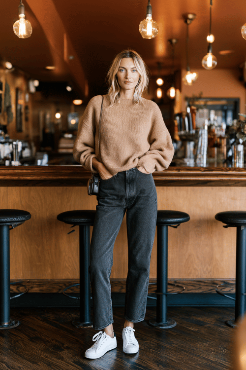 a woman wearing a cozy oversized camel sweater tucked into high-waisted dark wash straight-leg jeans, white sneakers, and a small crossbody bag