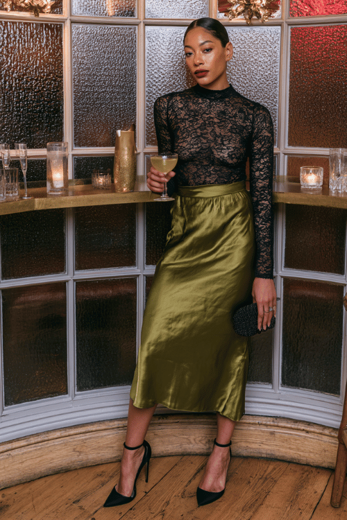 a woman wearing a dark olive green satin midi slip skirt and a fitted black lace long-sleeve top, black ankle strap heels and a small beaded clutch 