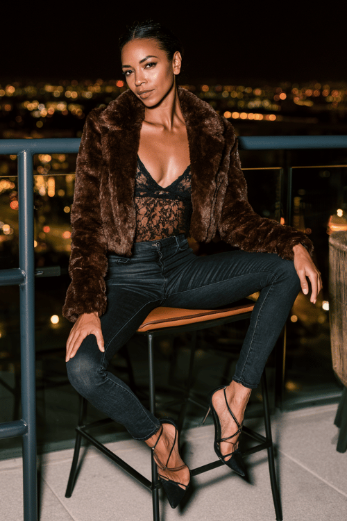 a woman wearing a luxurious deep chocolate brown faux fur cropped jacket over a black lace camisole, high-waisted dark skinny jeans, and black strappy stiletto heels 