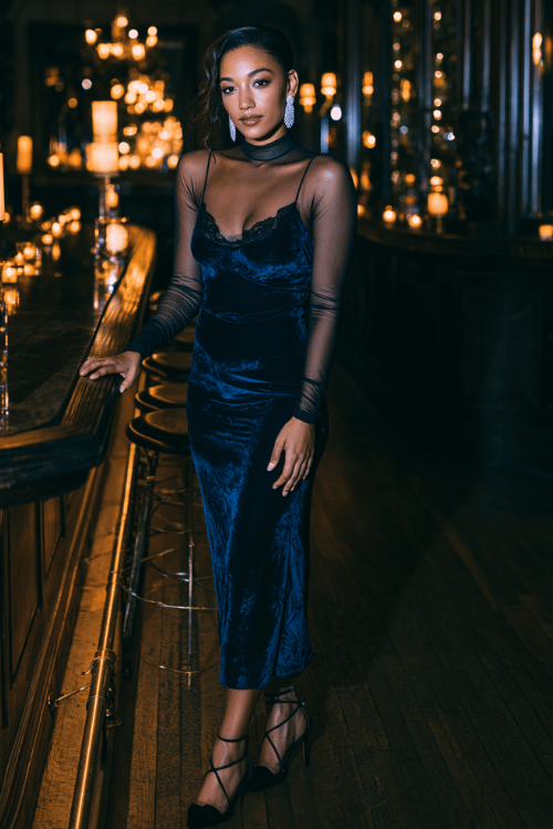 a woman wearing a luxurious deep navy blue velvet slip dress with spaghetti straps, layered over a long-sleeve black mesh top, black strappy heeled sandals and diamond drop earrings