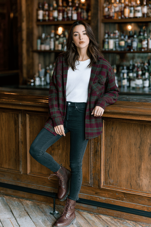 a woman wearing a plaid flannel shacket in burgundy and forest green over a white fitted long-sleeve top, skinny dark jeans, and brown lace-up ankle boots
