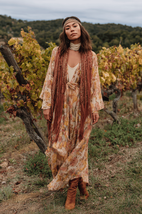 a woman wearing a romantic boho ensemble a sheer floral maxi dress in warm spice tones layered over a cream turtleneck, draped with a fringed blanket scarf 