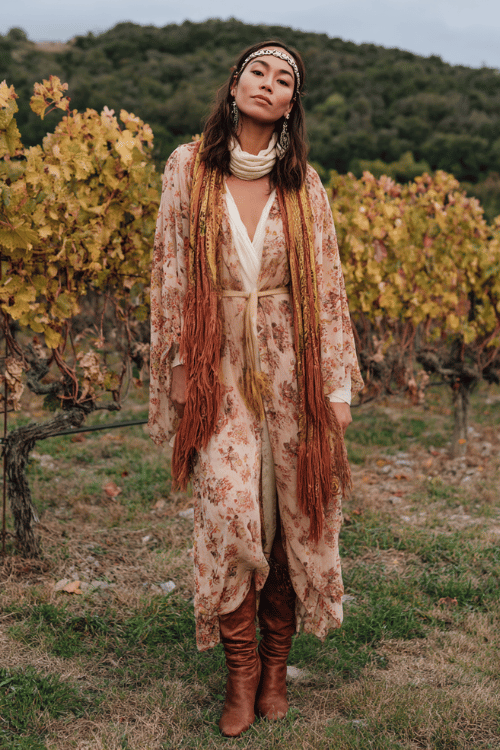 a woman wearing a romantic boho ensemble a sheer floral maxi dress in warm spice tones layered over a cream turtleneck, draped with a fringed blanket scarf