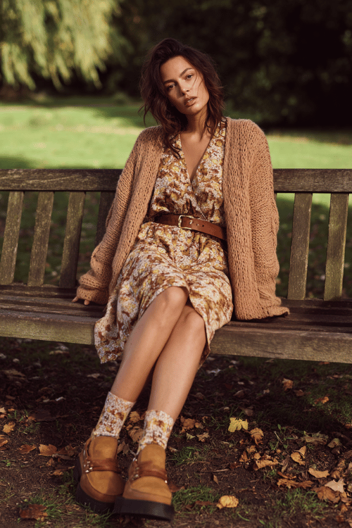 a woman wearing a soft oversized camel knit cardigan draped over a printed boho midi dress in muted autumn florals, cinched with a thin braided leather belt