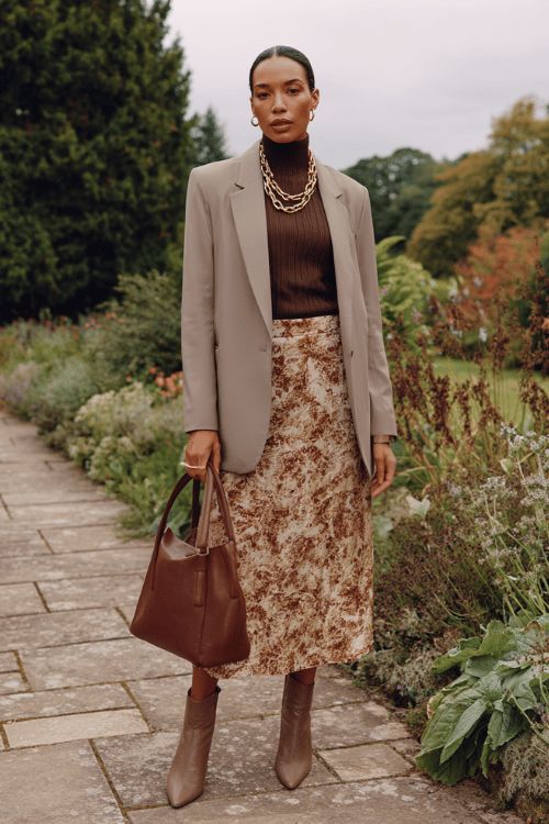 a woman wearing an elegant oversized longline blazer in warm stone grey over a flowing tiered midi skirt in soft earthy florals, paired with a fitted ribbed turtleneck