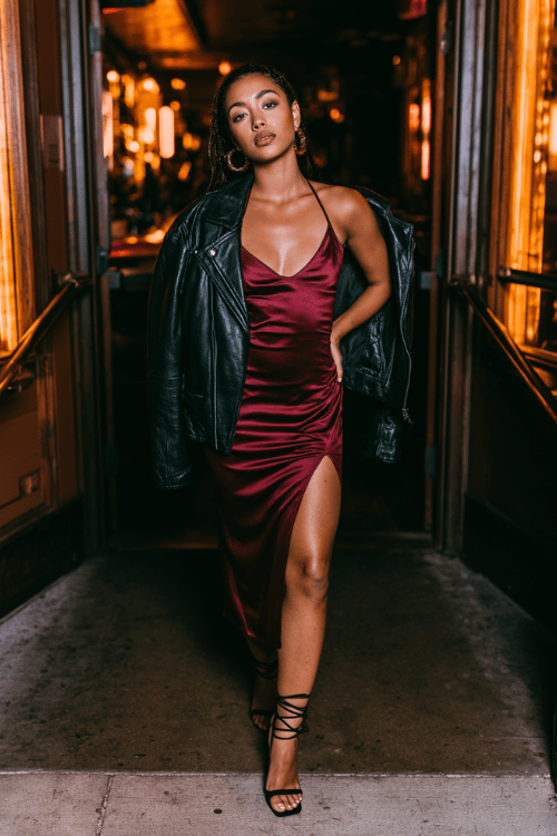 a young woman wearing a fitted burgundy satin slip dress with a black leather moto jacket draped over her shoulders, black strappy heeled sandals, gold hoop earrings
